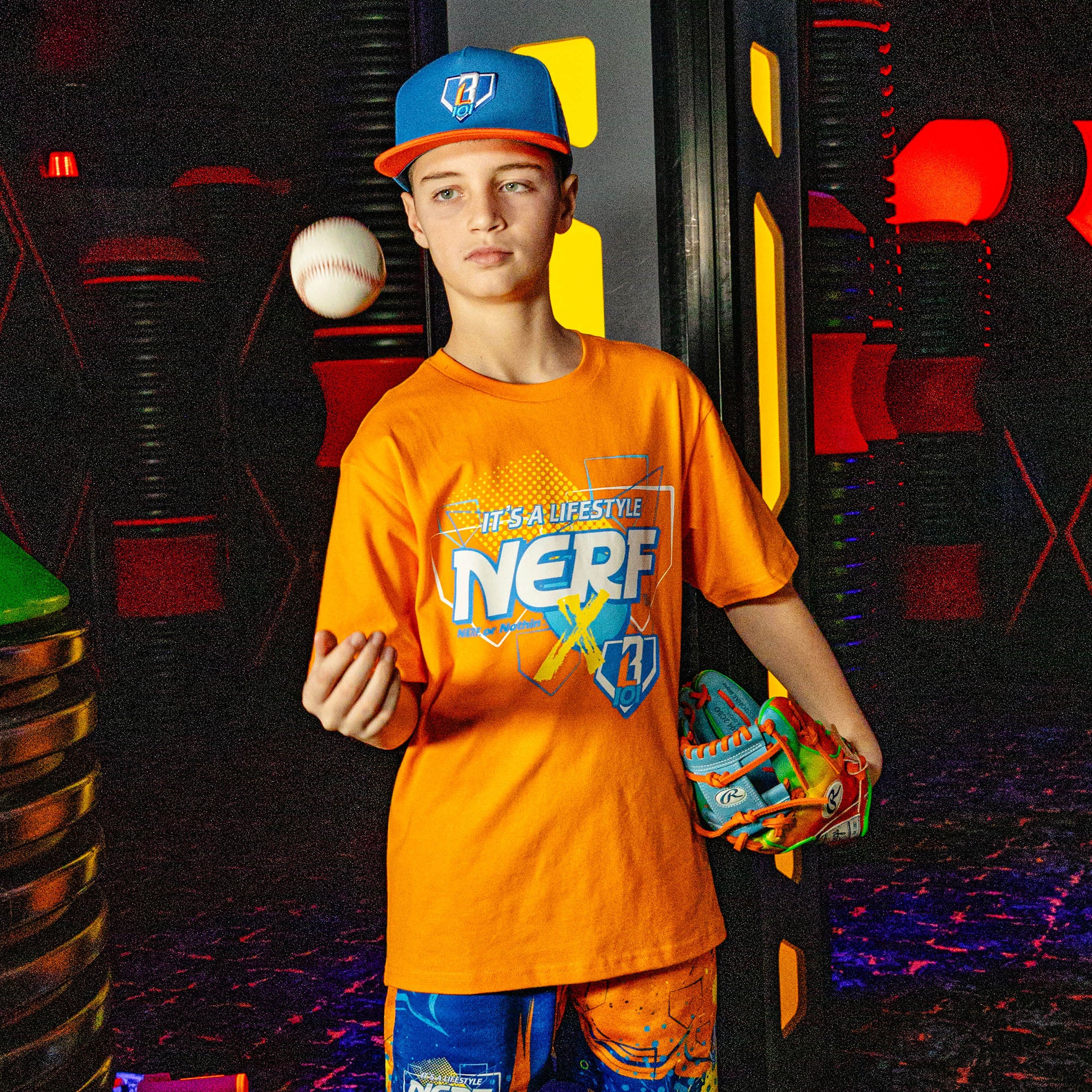 Person wearing an orange 'NERF X BL101' shirt holding a colorful baseball glove and ball in a colorful indoor setting.