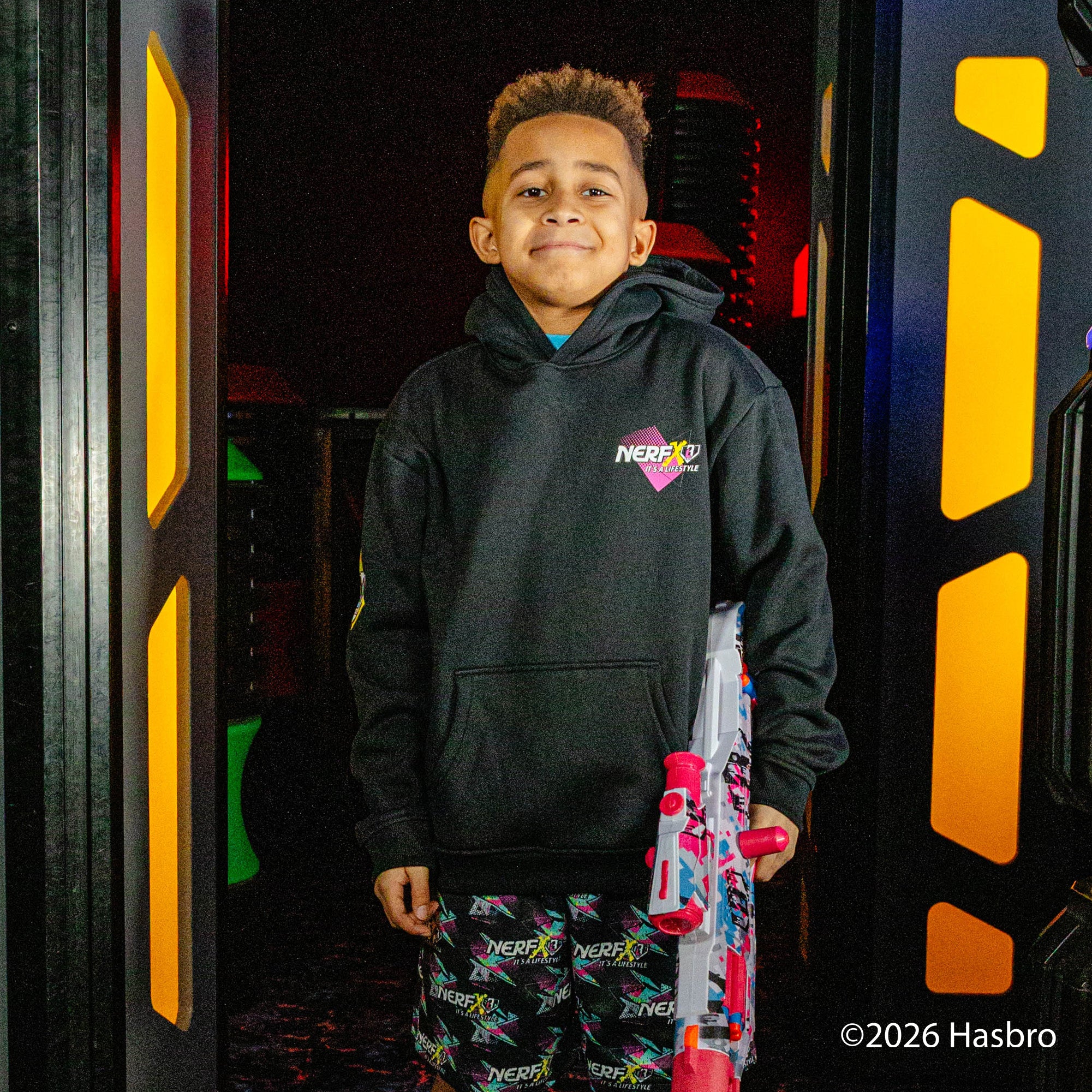 Child wearing a black hoodie with a 'Nerf X BL101' logo, holding a toy, in front of a colorful background.