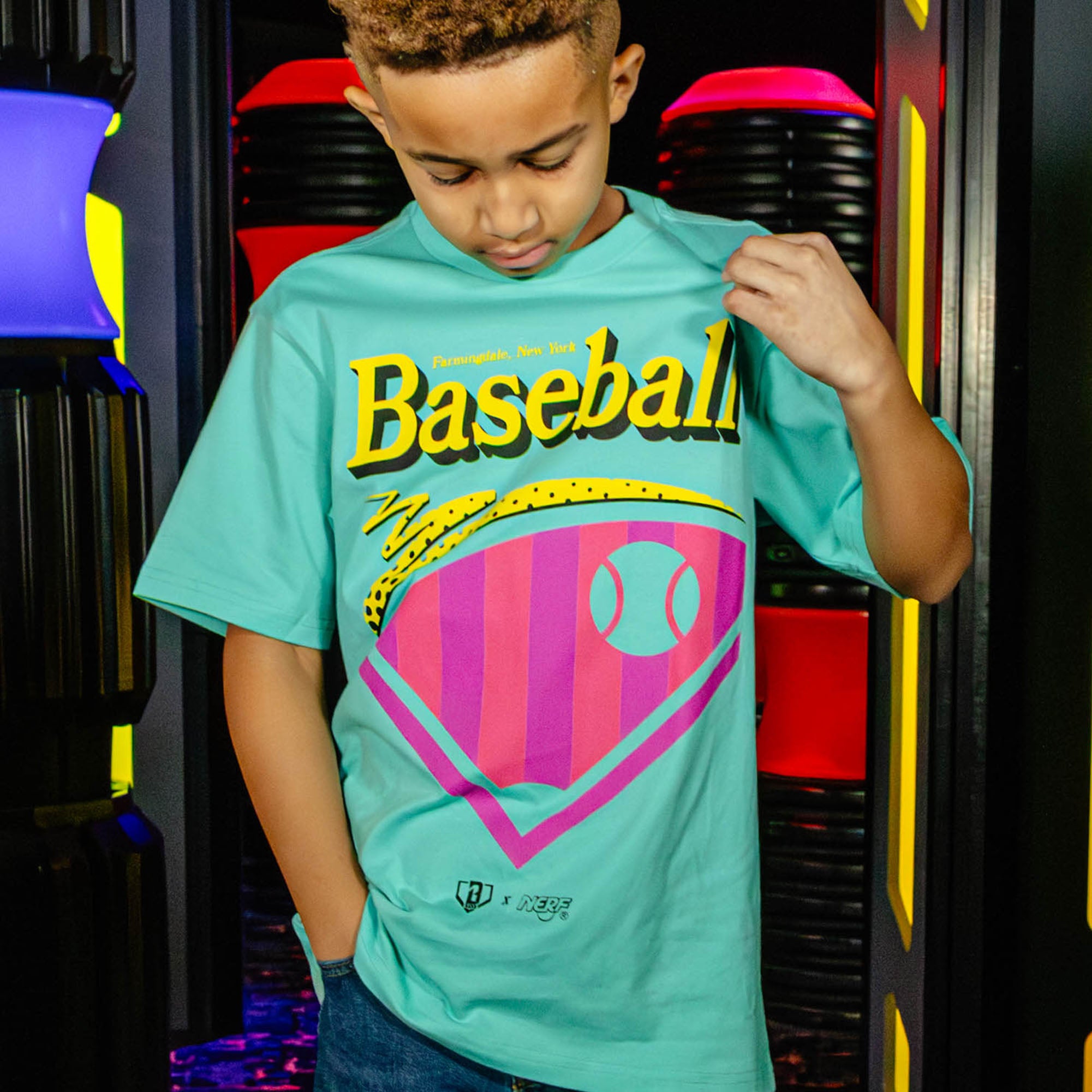 Child wearing a teal t-shirt with a colorful 'Baseball' design and 'BL101 X NERF' logo, standing in front of a colorful design.
