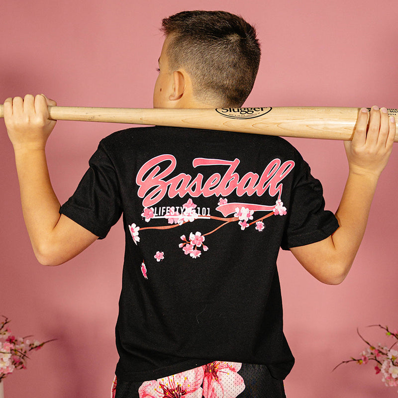 Young boy holding a wooden bat across his shoulders showing the back of a black BL101 tee with pink "Baseball" script and cherry blossom graphic against a pink background