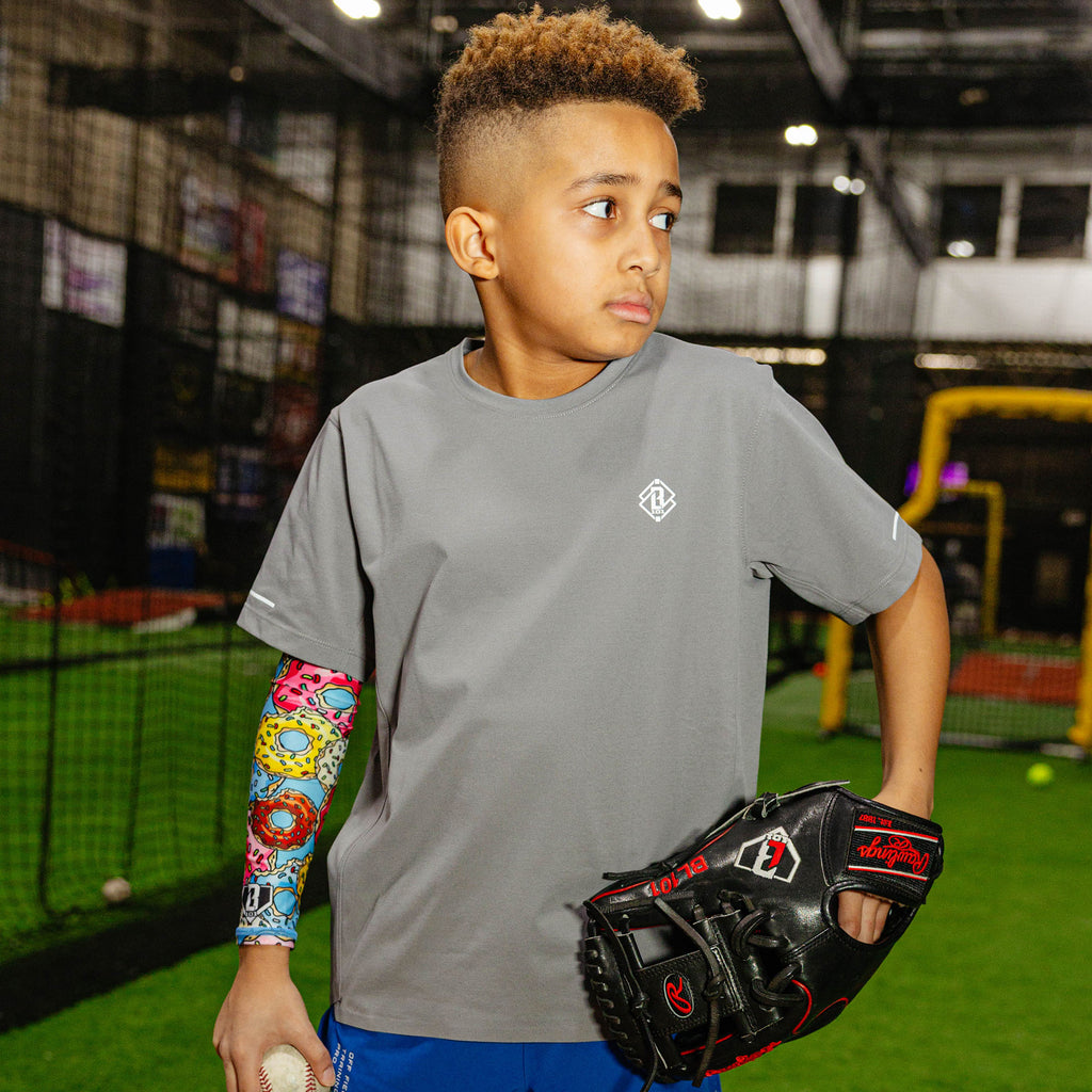 Young baseball player in a gray BL101 shirt wearing the donut arm sleeve on his left arm while holding a glove and baseball in an indoor facility