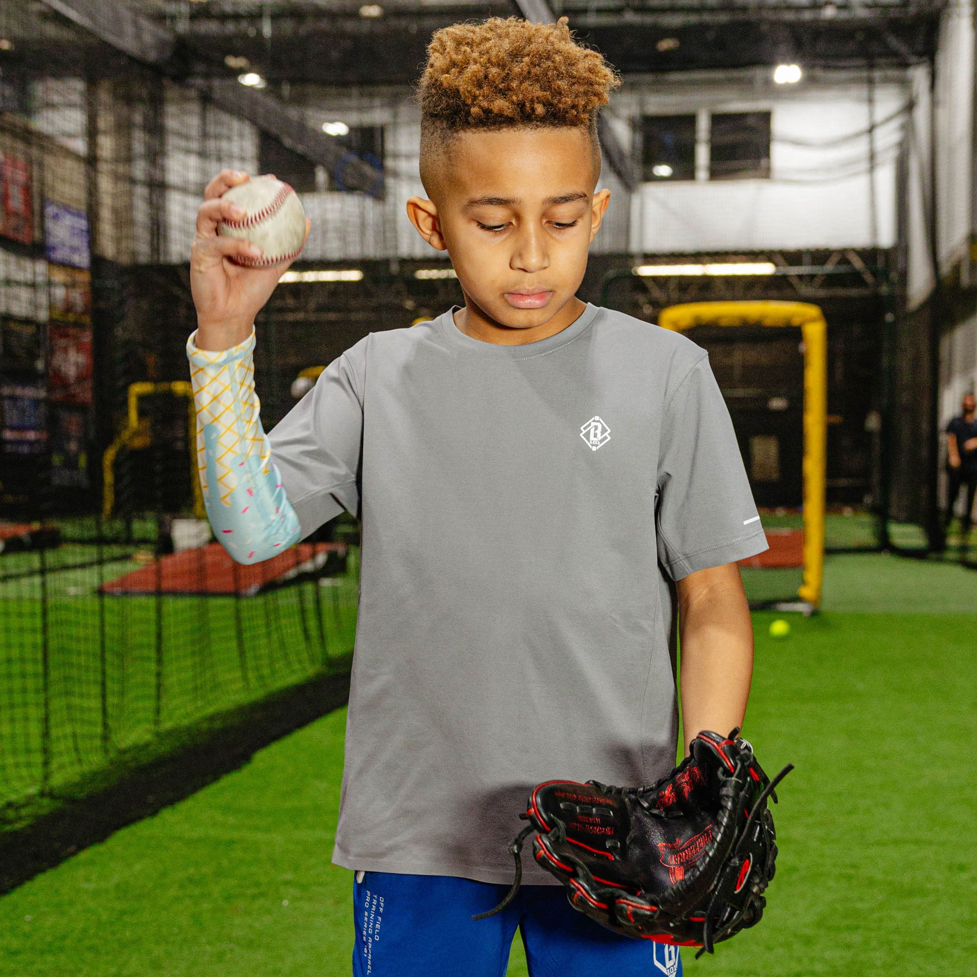 Young baseball player in a gray BL101 shirt holding up a baseball and a glove while wearing the cotton candy ice cream arm sleeve in an indoor facility