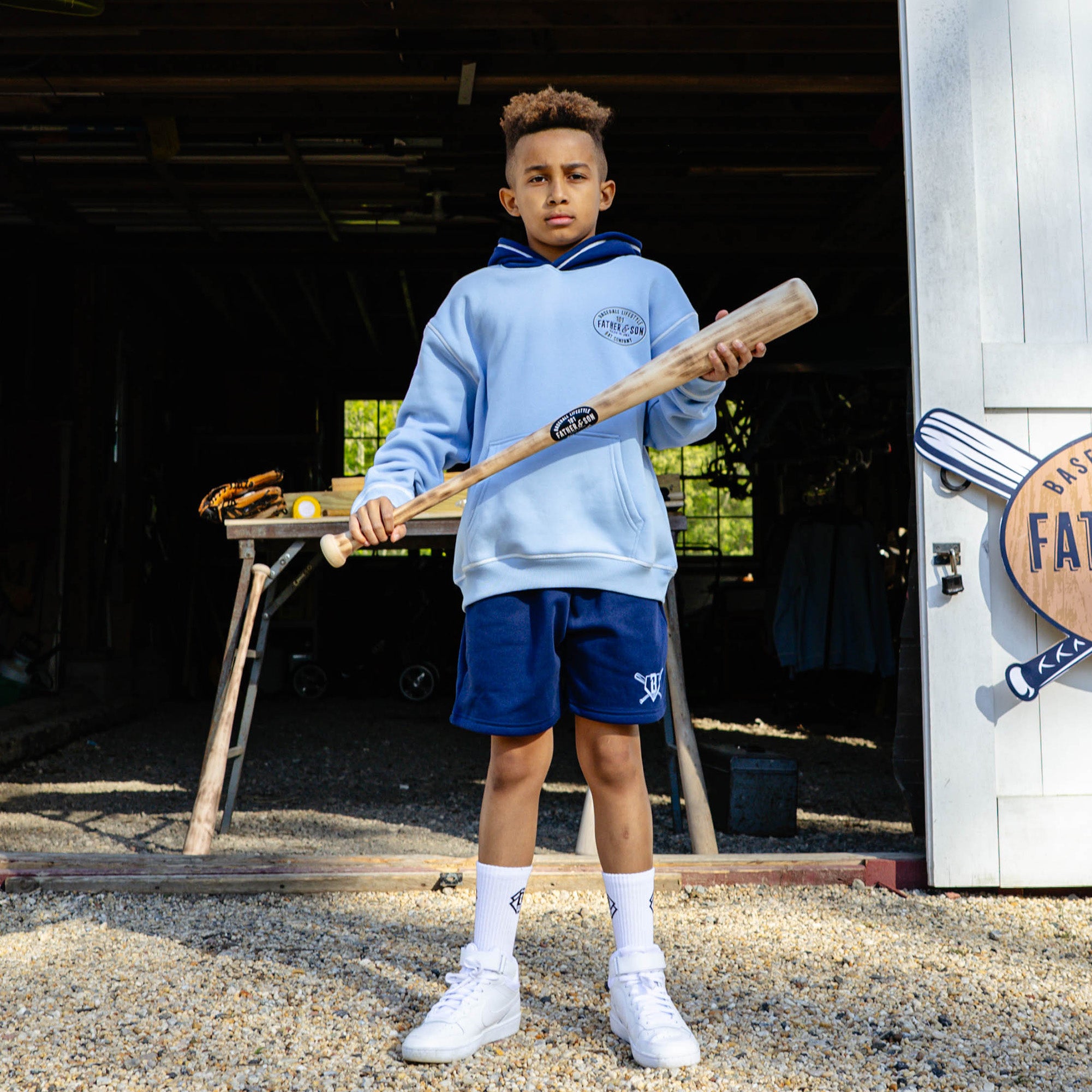Young boy holding a wooden baseball bat and wearing the columbia blue youth BL101 Bat Co Hoodie with navy contrast hood, paired with navy Heritage Diamond Sweatshorts and white BL101 crew socks.