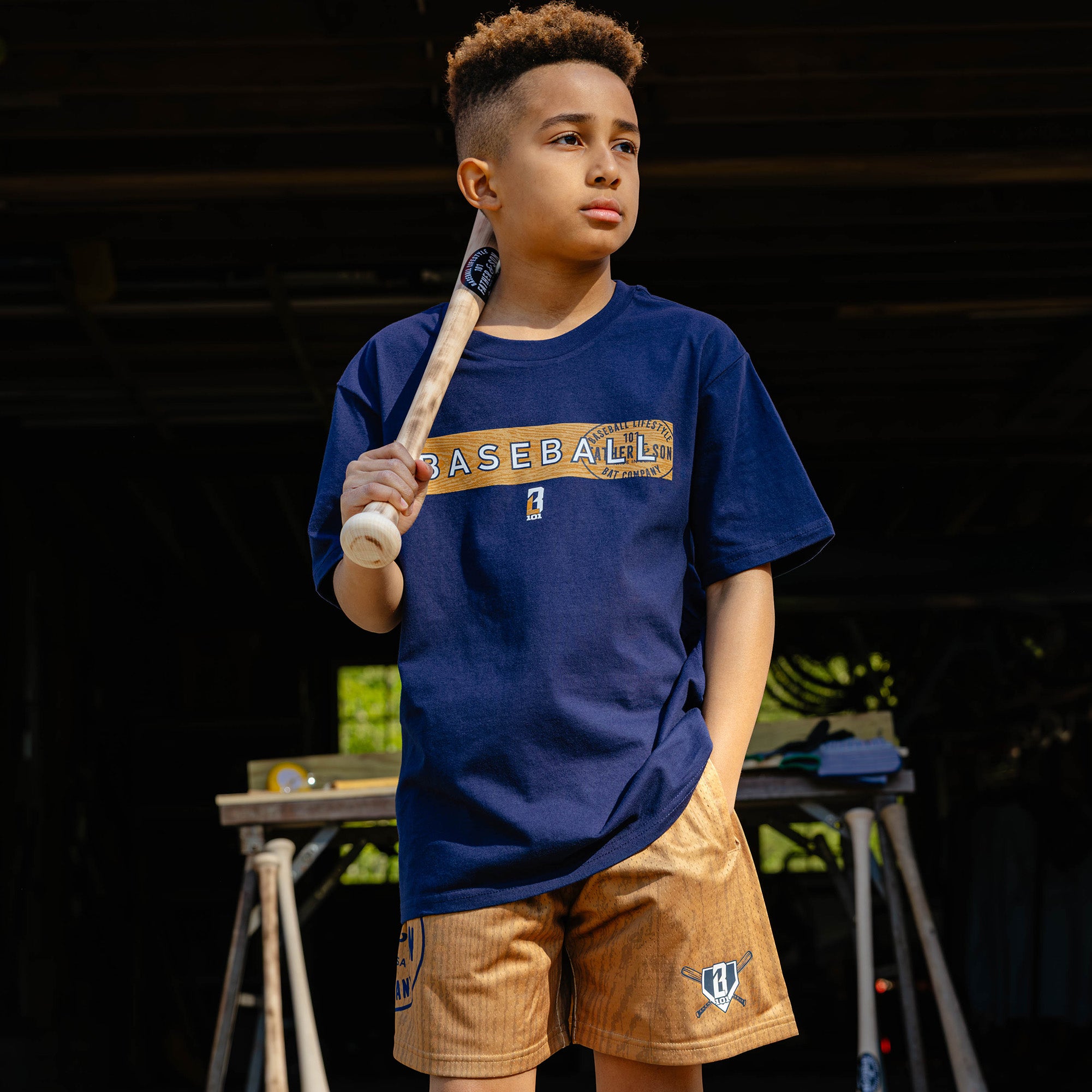 Young boy resting a baseball bat on his shoulder wearing the brown youth BL101 Bat Co Shorts paired with the navy Radar Tee.