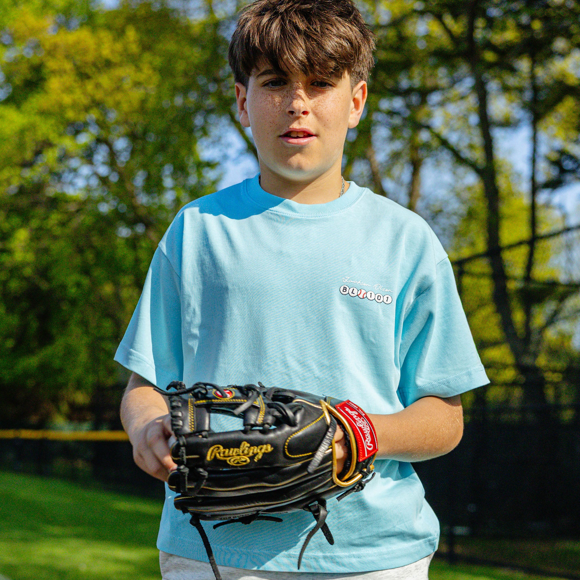Youth player holding a baseball glove on a field, wearing the light blue Jackson Olson x BL101 BSBL Era relaxed tee with chest logo