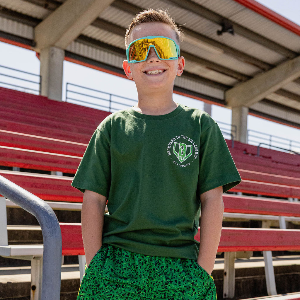 Smiling boy at red stadium bleachers wearing teal shield sunglasses a forest green tee with a left chest BL101 logo graphic and green printed shorts