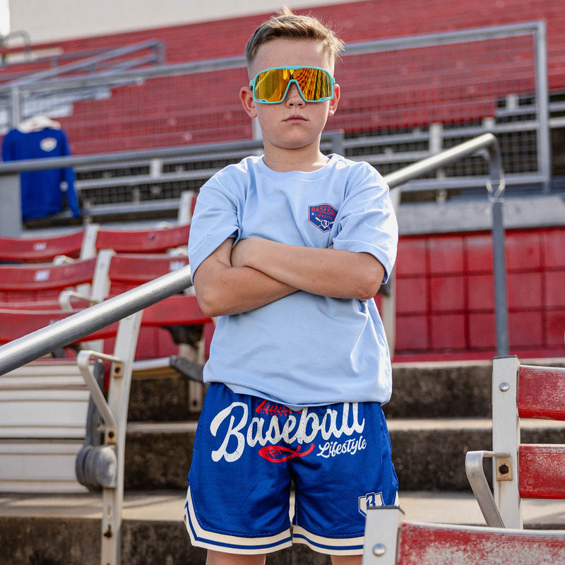Boy with arms crossed at red stadium bleachers wearing teal shield sunglasses a light blue tee with a left chest BL101 logo graphic and royal blue Baseball Lifestyle mesh shorts