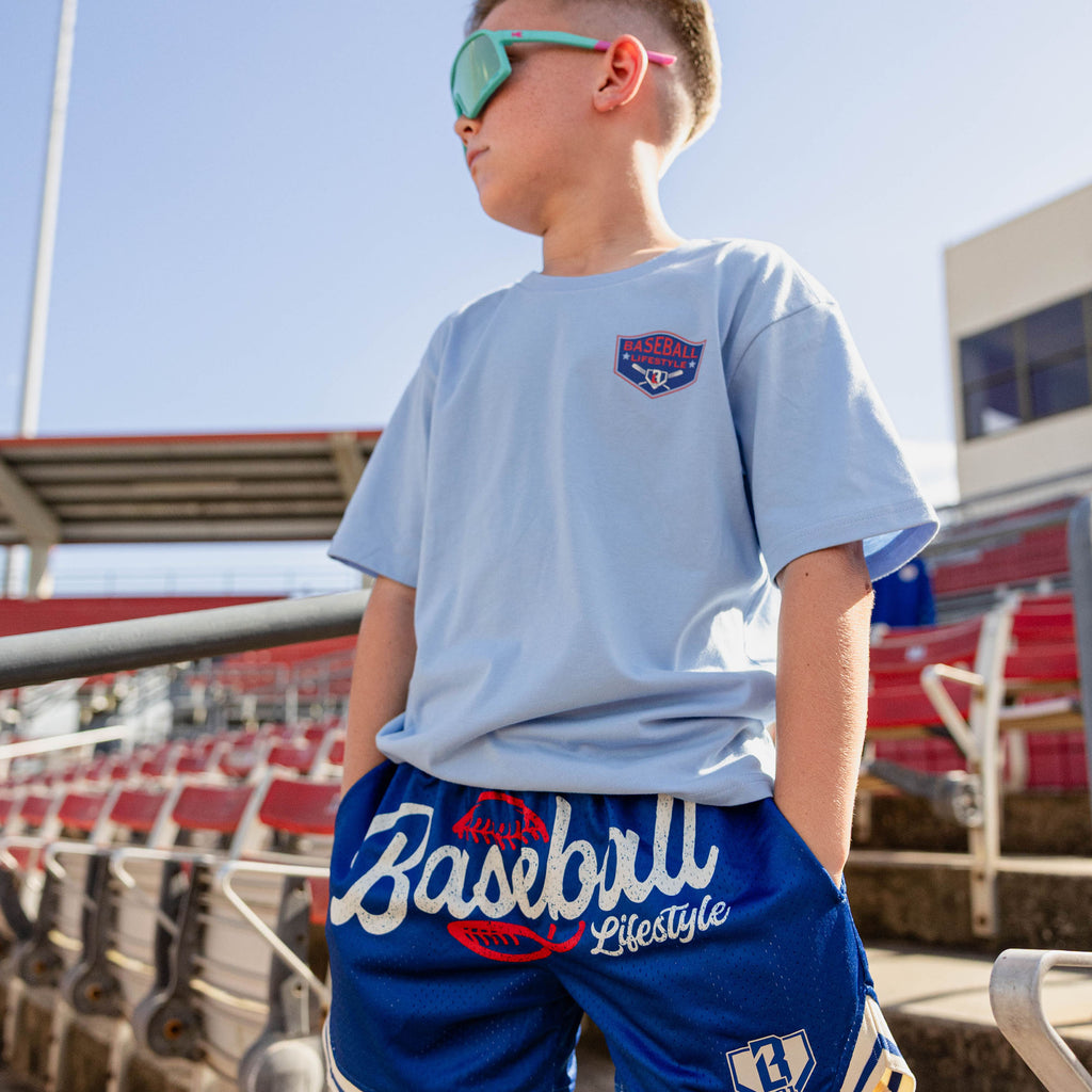Boy looking up at red stadium bleachers wearing teal shield sunglasses a light blue Baseball Lifestyle shield tee and royal blue Baseball Lifestyle mesh shorts