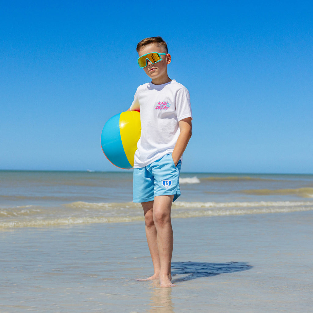 Young boy on the beach wearing light blue BL101 youth swim trunks and a white Rain Delay tee, holding a beach ball while wearing yellow mirrored shield sunglasses.