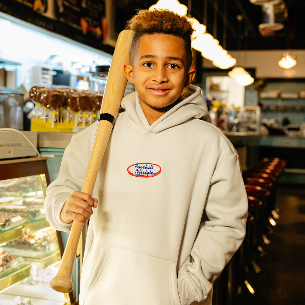 Child wearing a beige hoodie with 'Baseball Lifestyle' logo holding a baseball bat in a diner