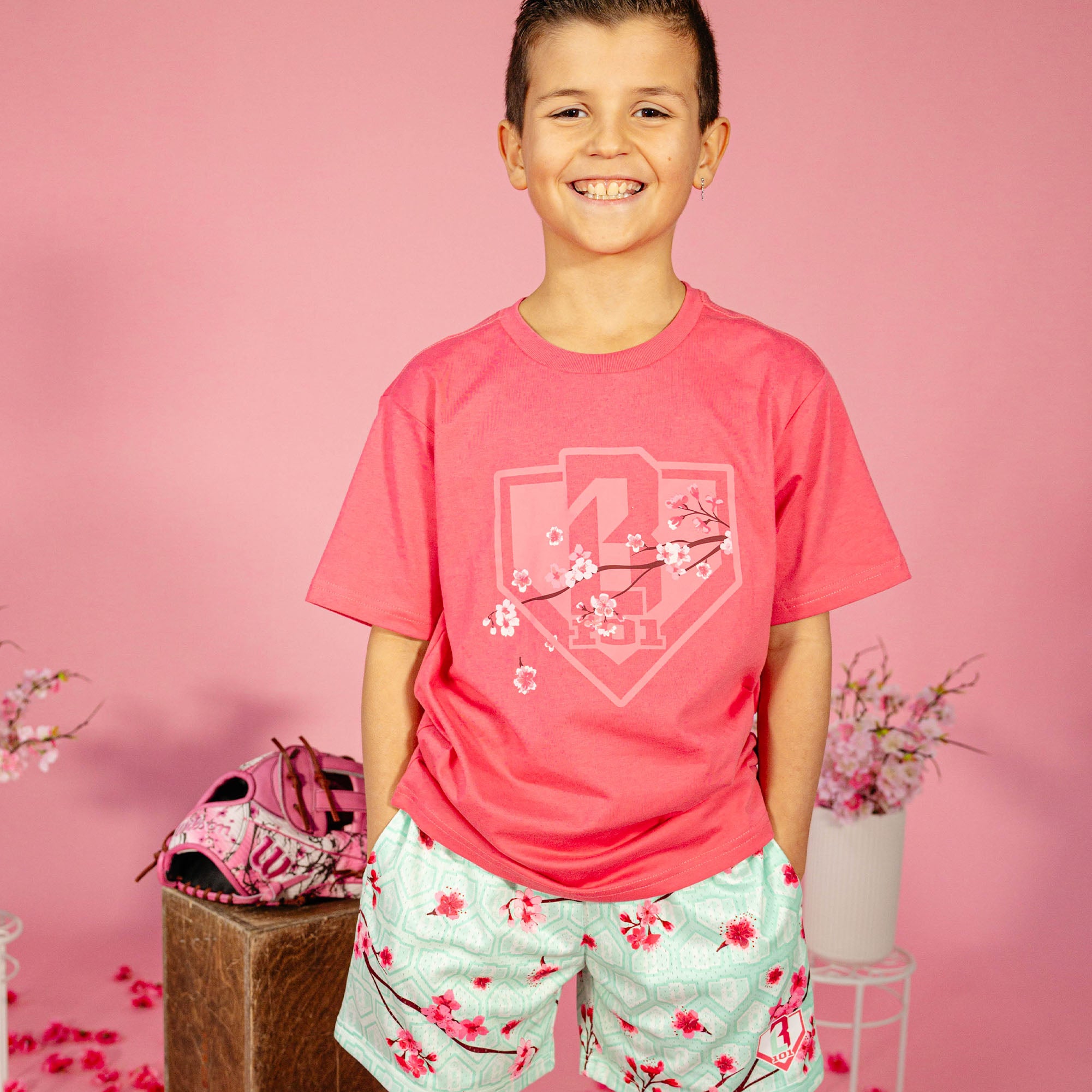 Young boy wearing a pink BL101 cherry blossom shield tee and mint green BL101 cherry blossom mesh shorts against a pink background, front view