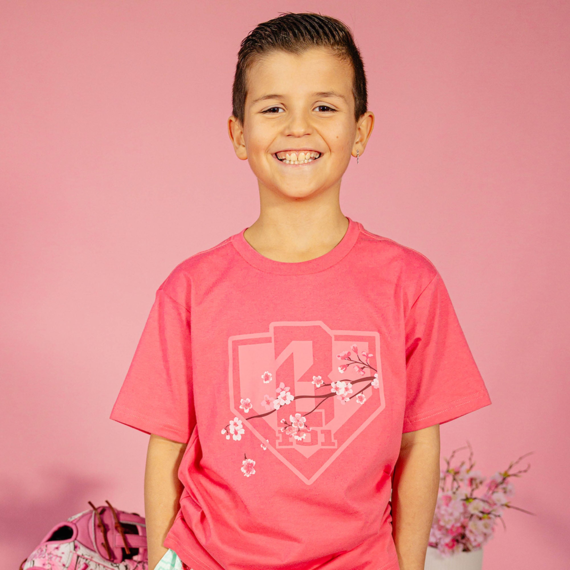 Young boy wearing a pink BL101 cherry blossom shield tee against a pink background, front view