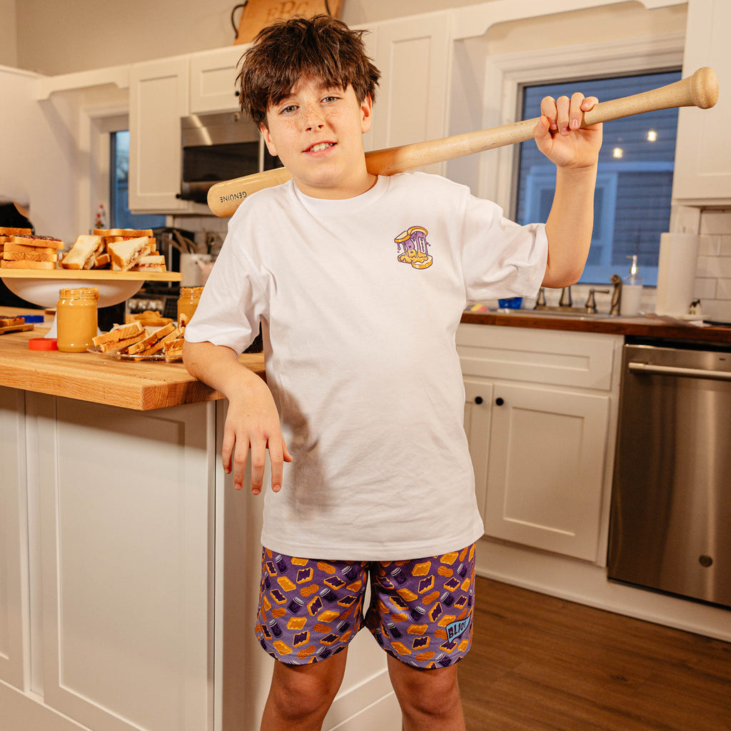 Young boy holding a baseball bat in a kitchen, wearing BL101 youth purple PB&J print mesh shorts and a white BL101 tee.
