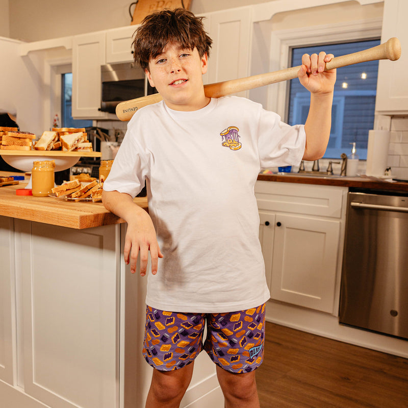 Young boy holding a baseball bat in a kitchen, wearing BL101 youth purple PB&J print mesh shorts and a white BL101 tee.