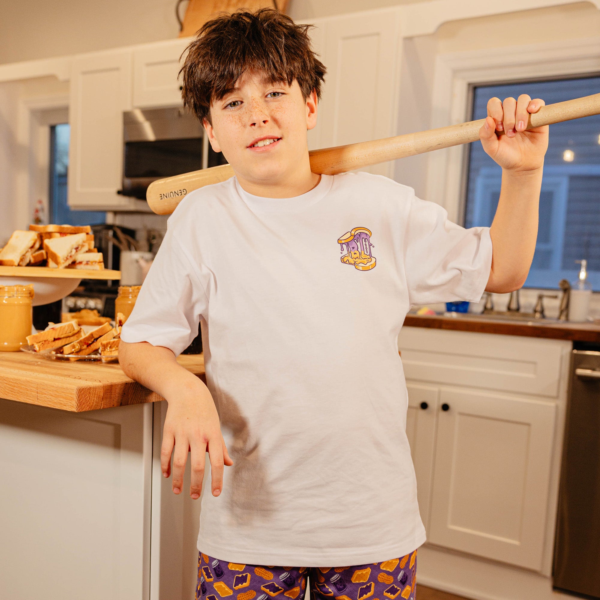 Young boy holding a baseball bat in a kitchen wearing the BL101 youth Peanut Butter and Jelly white tee, showing the chest logo.
