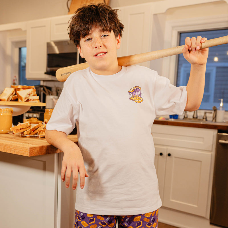 Young boy holding a baseball bat in a kitchen wearing the BL101 youth Peanut Butter and Jelly white tee, showing the chest logo.