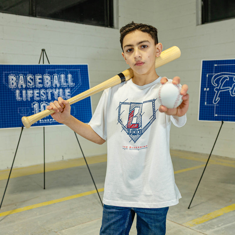 Person wearing a white t-shirt with 'The Blueprint' text, blue and red BL101 logo while holding a baseball bat and ball 