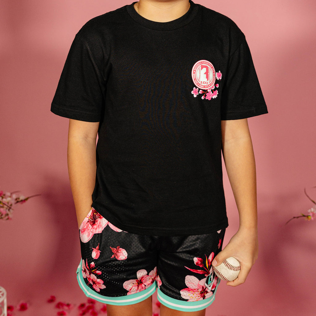 Young boy wearing a black BL101 cherry blossom tee and black BL101 cherry blossom mesh shorts with teal hem while holding a baseball against a pink background, front view