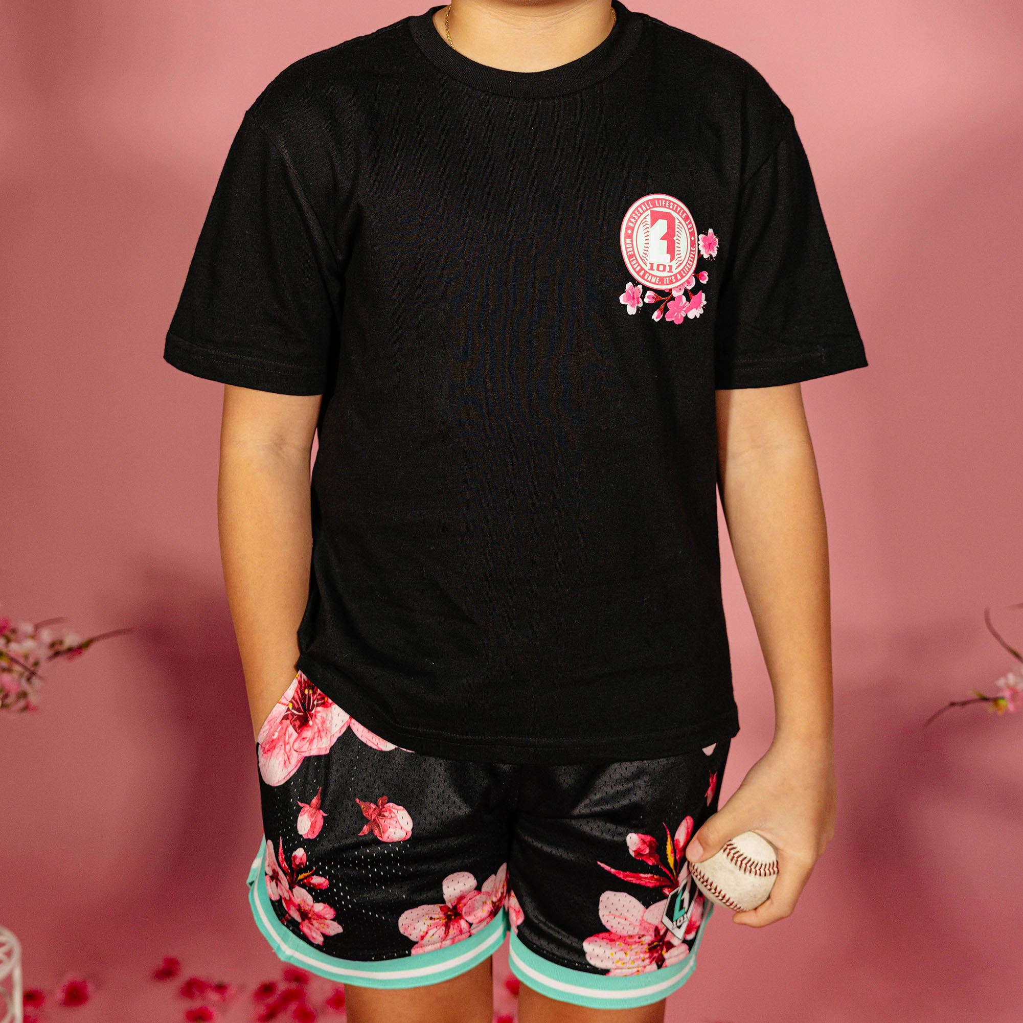 Young boy wearing a black BL101 cherry blossom tee and black BL101 cherry blossom mesh shorts with teal hem while holding a baseball against a pink background, front view