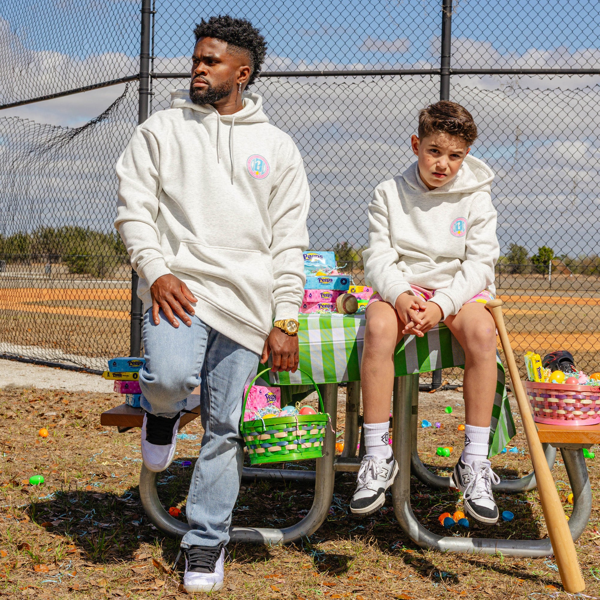 Two wearing grey hoodie with colorful BL101 logo, sitting on a playground swing set with colorful toys around them.
