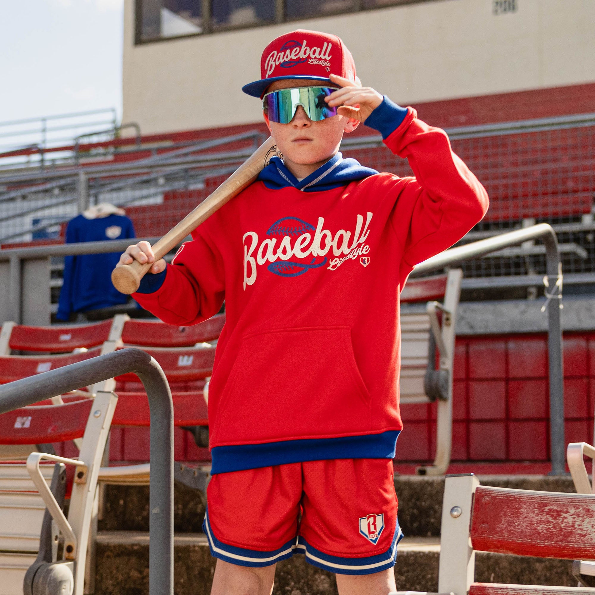 Boy holding a bat on his shoulder at red stadium bleachers wearing iridescent shield sunglasses a red Baseball Lifestyle snapback and a red Baseball Lifestyle hoodie with red mesh shorts
