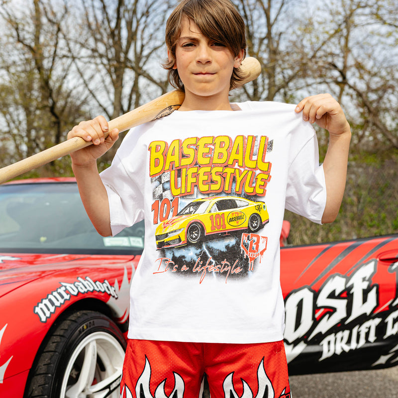 Young boy holding wooden bat across shoulders, wearing white Diamond Drift tee with yellow and red BL101 race car graphic and red flame shorts, in front of wrapped red car.