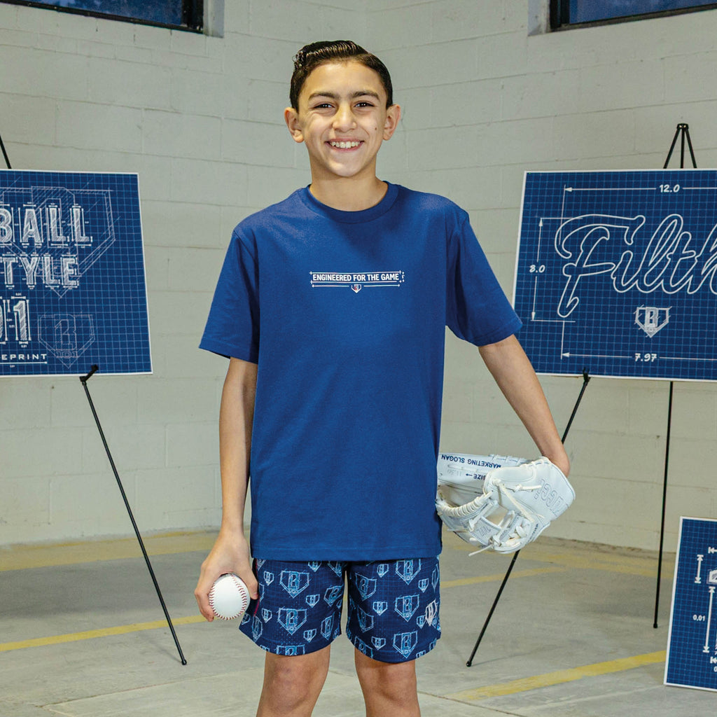 Young boy in blue t-shirt with a BL101 logo and "Engineered for the game" text, standing in front of sports-themed wall decorations.