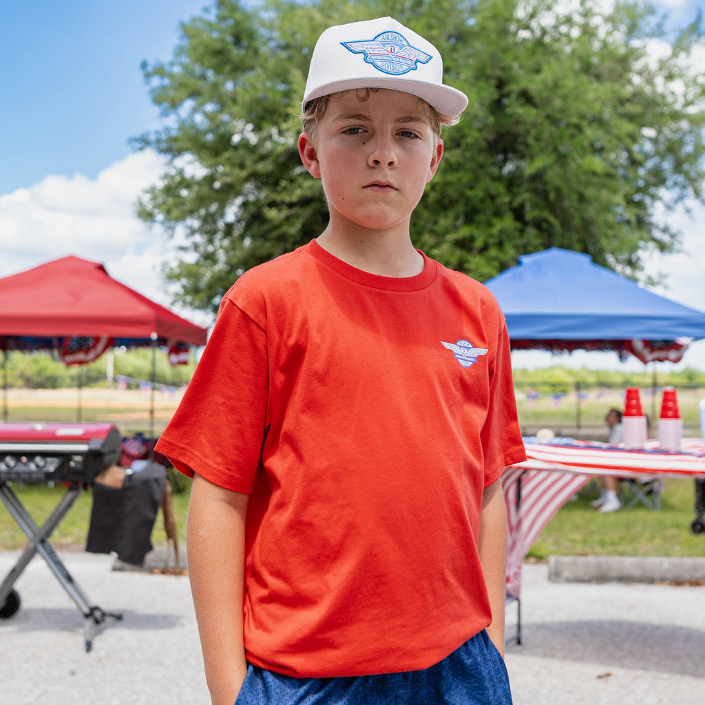 Front-facing youth male model wearing gray Air Show National Air Races Division snapback and orange tee outdoors at baseball event