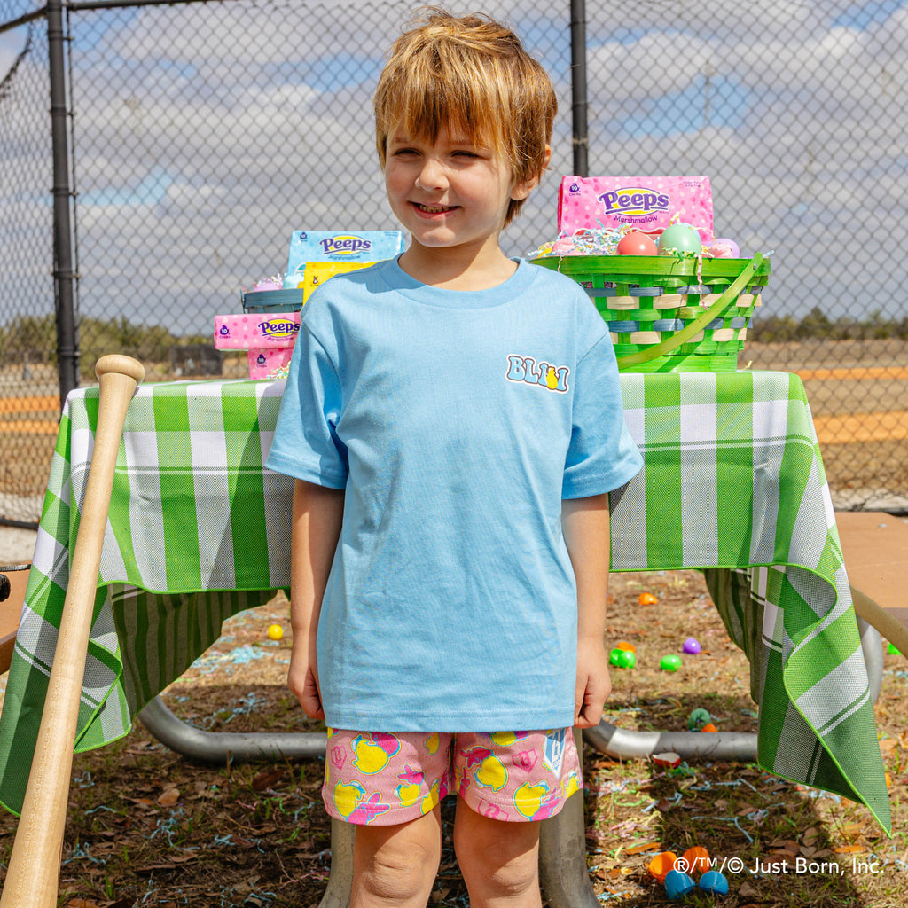 Child standing in front of a table with Easter baskets and eggs, wearing a blue peeps BL101 shirt and colorful shorts.