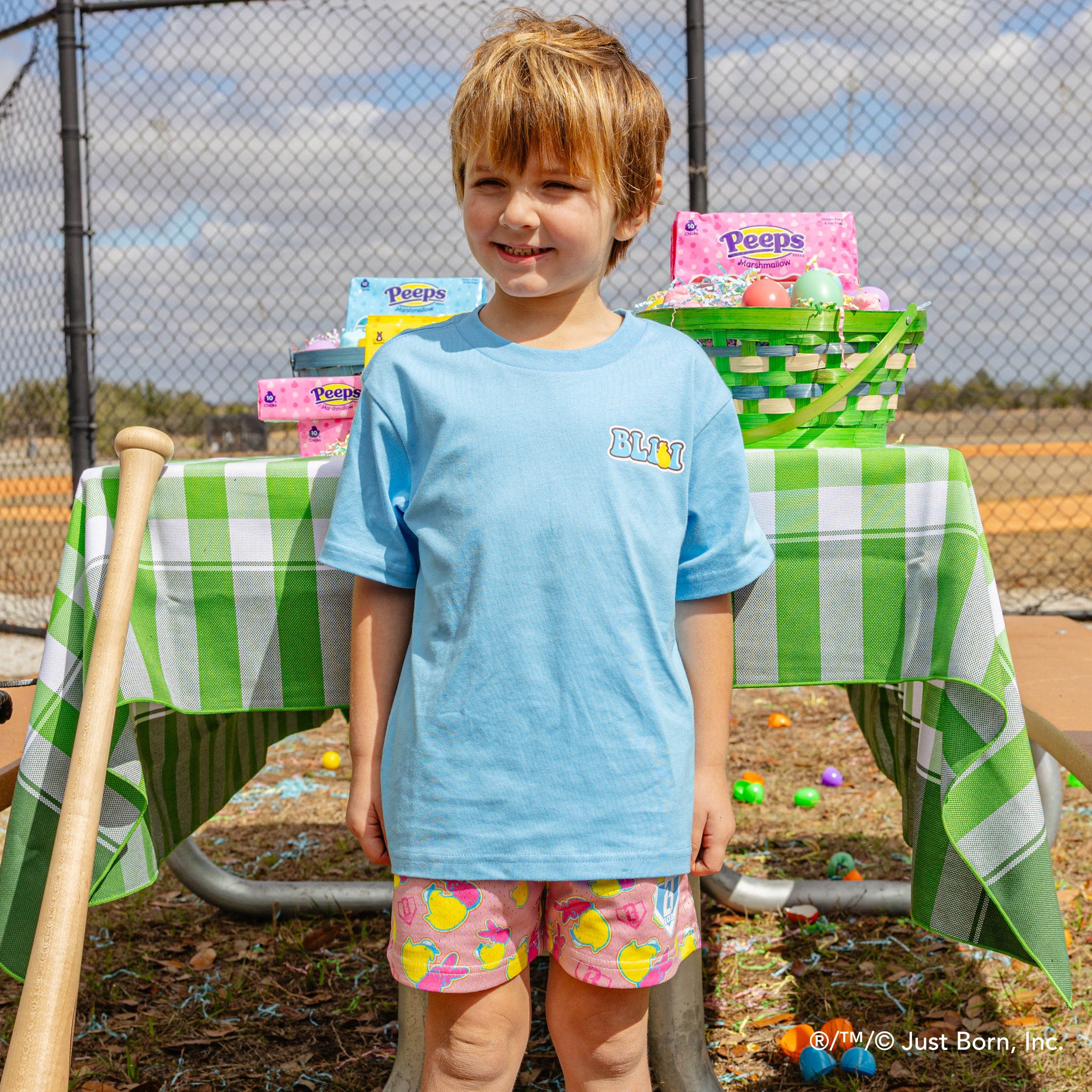 Child standing in front of a table with Easter baskets and eggs, wearing a blue peeps BL101 shirt and colorful shorts.