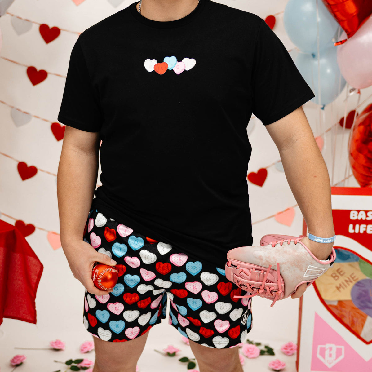 Person wearing a black t-shirt with colorful heart design and heart patterned shorts in front of a decorative background.