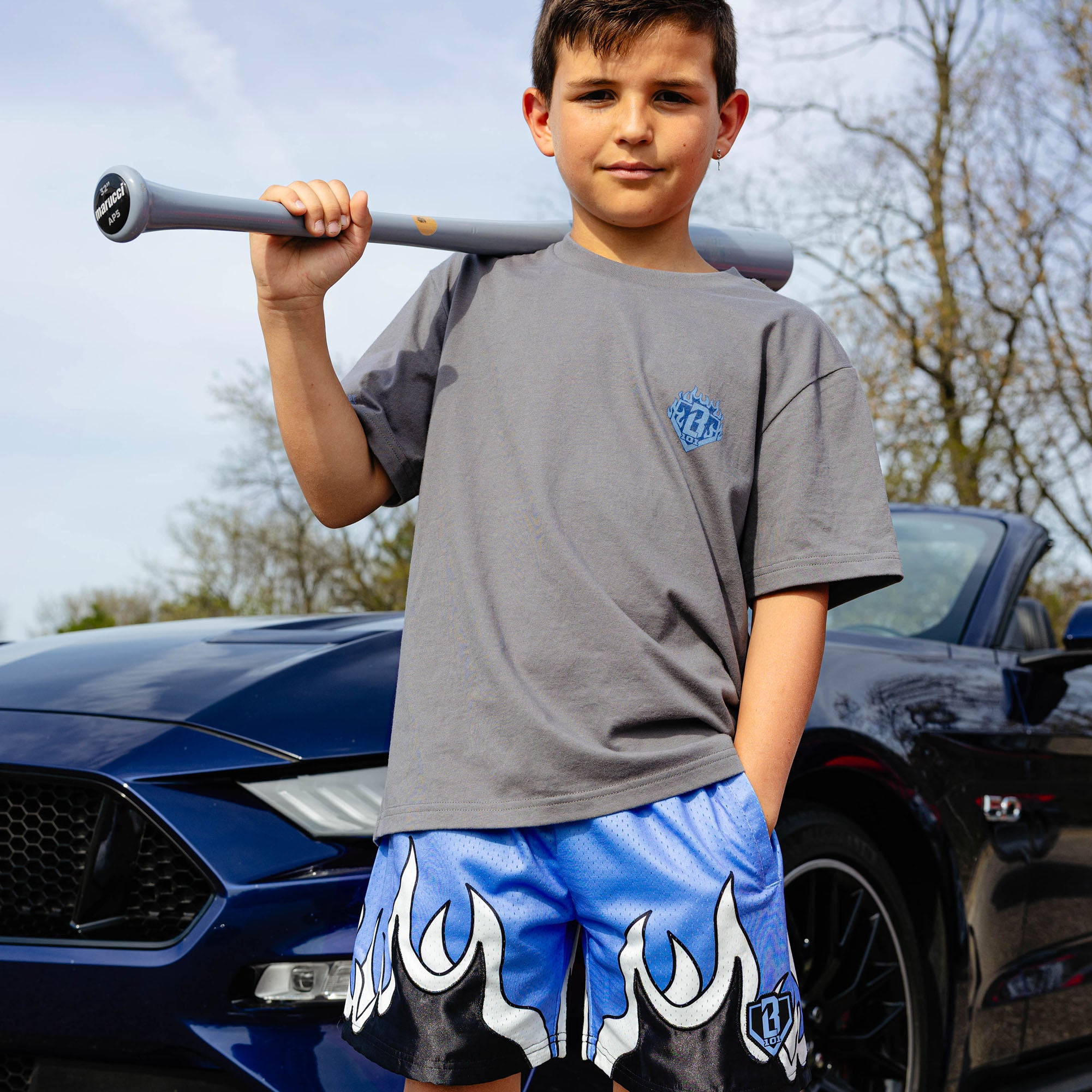 Young boy holding gray baseball bat over shoulder, wearing gray Flame Thrower tee and blue flame mesh shorts, standing in front of navy Ford Mustang.