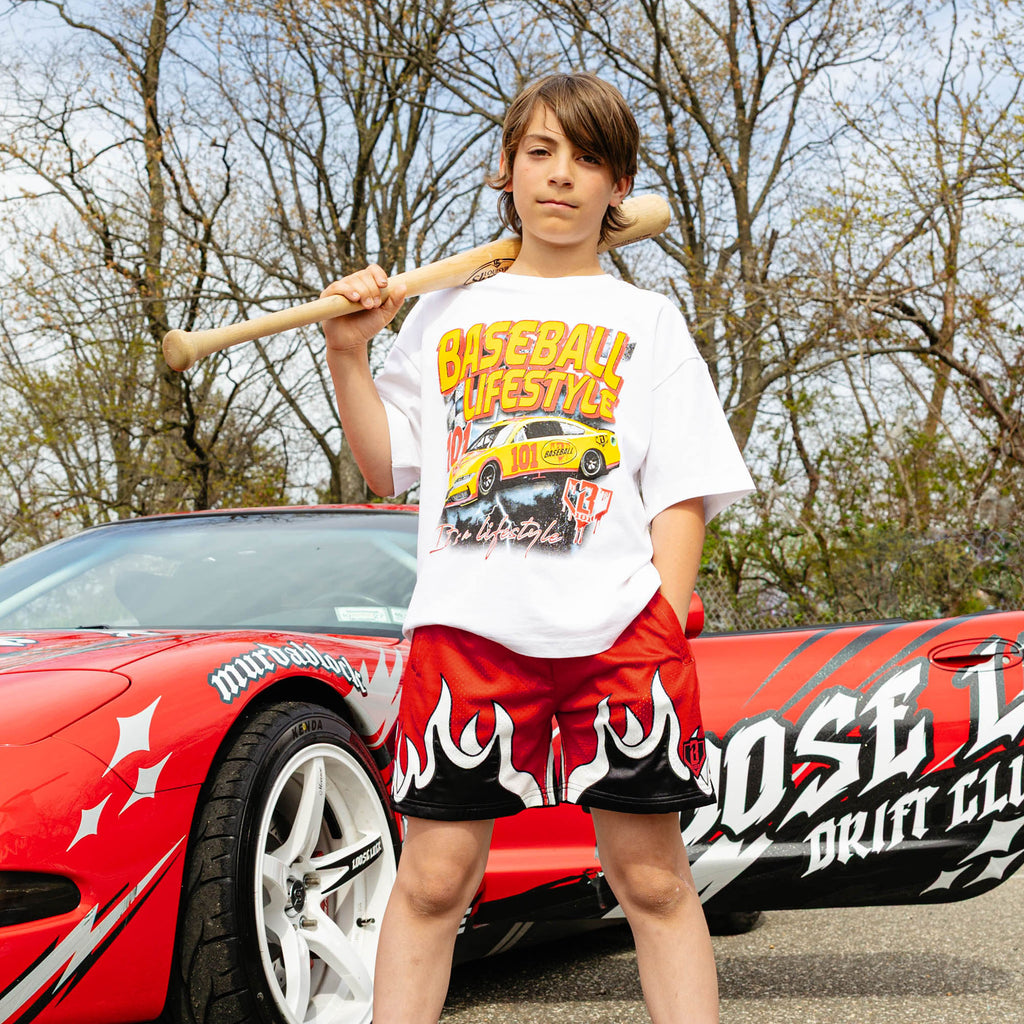 Young boy holding wooden bat over shoulder, wearing white BL101 race car tee and red flame mesh shorts, standing beside wrapped red drift car.