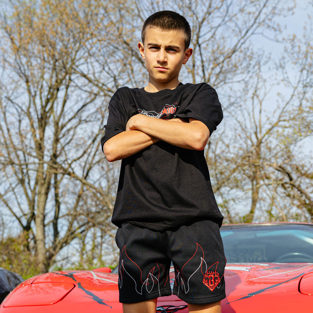 Young boy standing on red Corvette hood arms crossed, wearing black Flames Stitched tee and matching black flame sweatshorts.