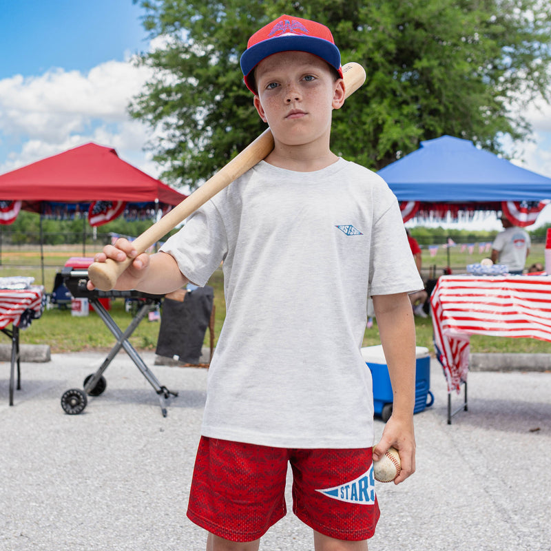 Front-facing youth male model wearing red Freedom Swings shorts and heather gray tee holding baseball bat and ball at outdoor baseball event