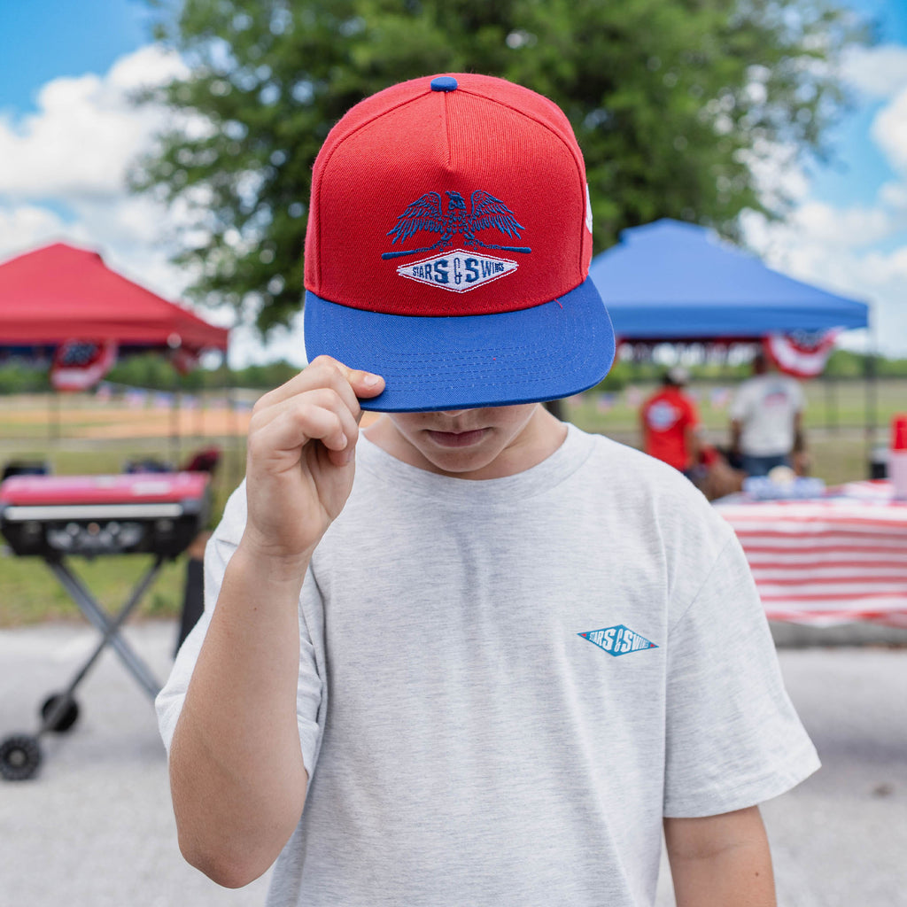 Person wearing a red and blue cap with a Freedom Swings logo, standing outdoors with tents and people in the background.
