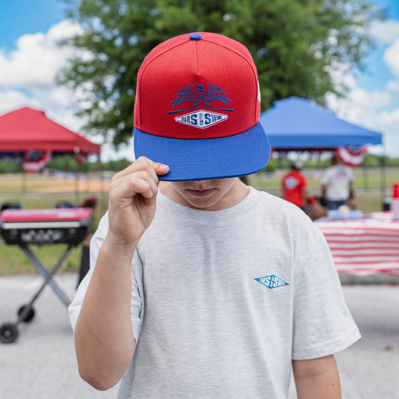 Person wearing a red and blue cap with a Freedom Swings logo, standing outdoors with tents and people in the background.