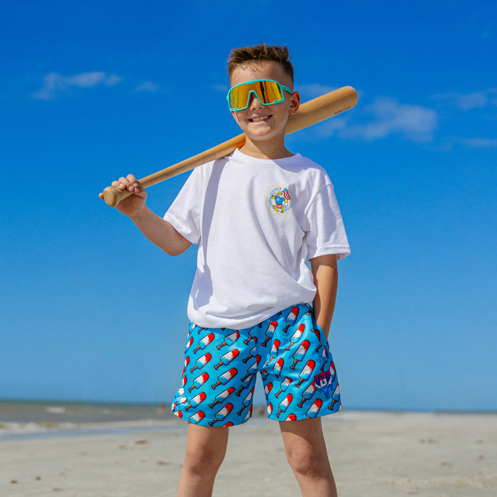 Young boy on the beach wearing BL101 youth popsicle bat print swim trunks, white BL101 tee, and yellow mirrored shield sunglasses, resting a bat on his shoulder.