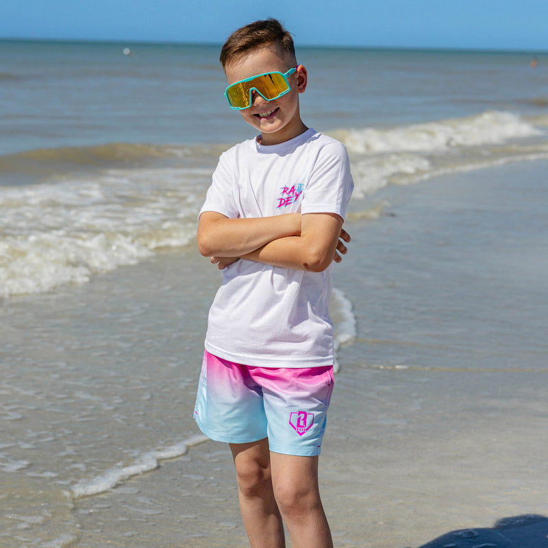 Young boy on the beach wearing BL101 youth pink to blue ombre swim trunks and a white Rain Delay tee, arms crossed and smiling with yellow mirrored shield sunglasses.