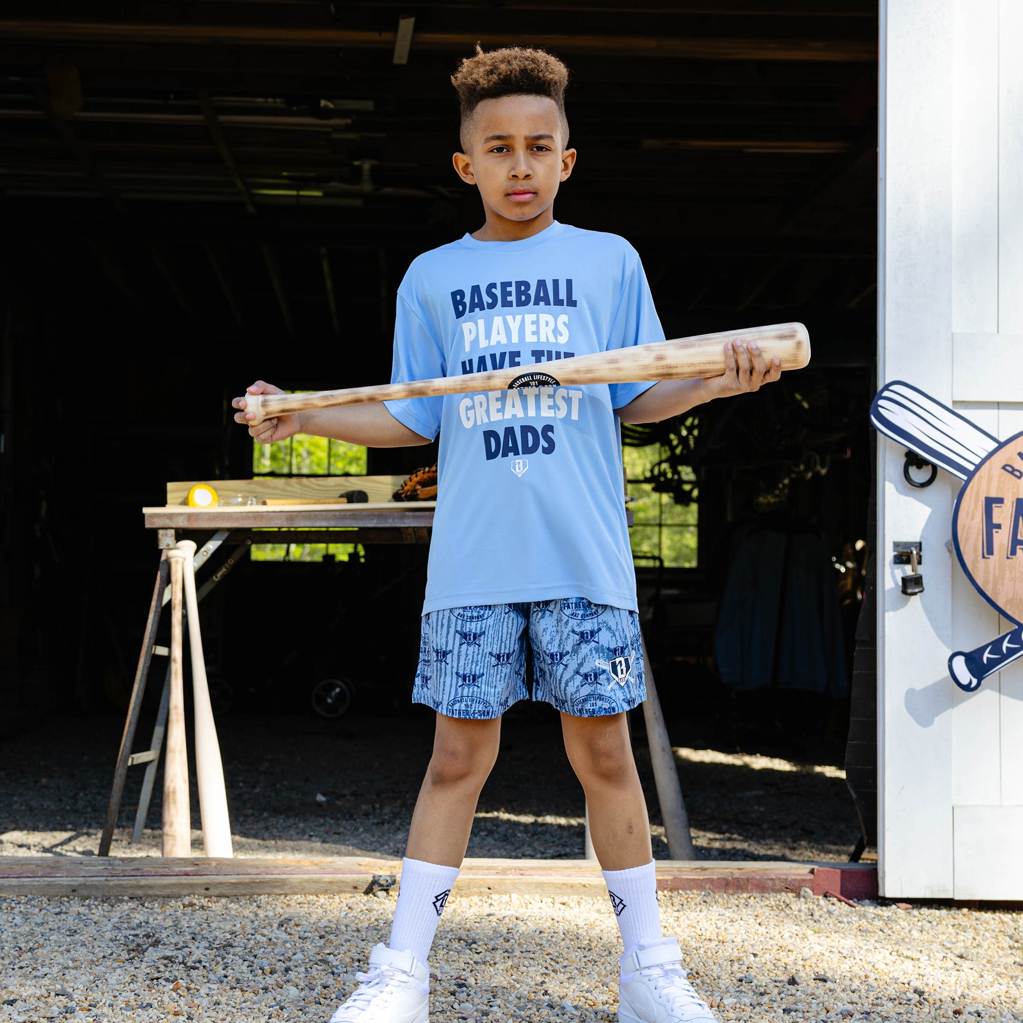 Young boy holding a wooden baseball bat horizontally in front of him while wearing the columbia blue youth Heritage Diamond Shorts paired with the columbia blue Greatest Dad Athletic Tee and white BL101 crew socks.