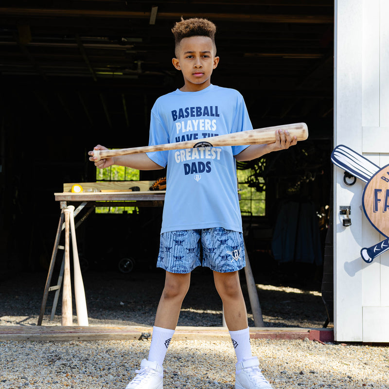 Young boy holding a wooden baseball bat horizontally in front of him while wearing the columbia blue youth Heritage Diamond Shorts paired with the columbia blue Greatest Dad Athletic Tee and white BL101 crew socks.