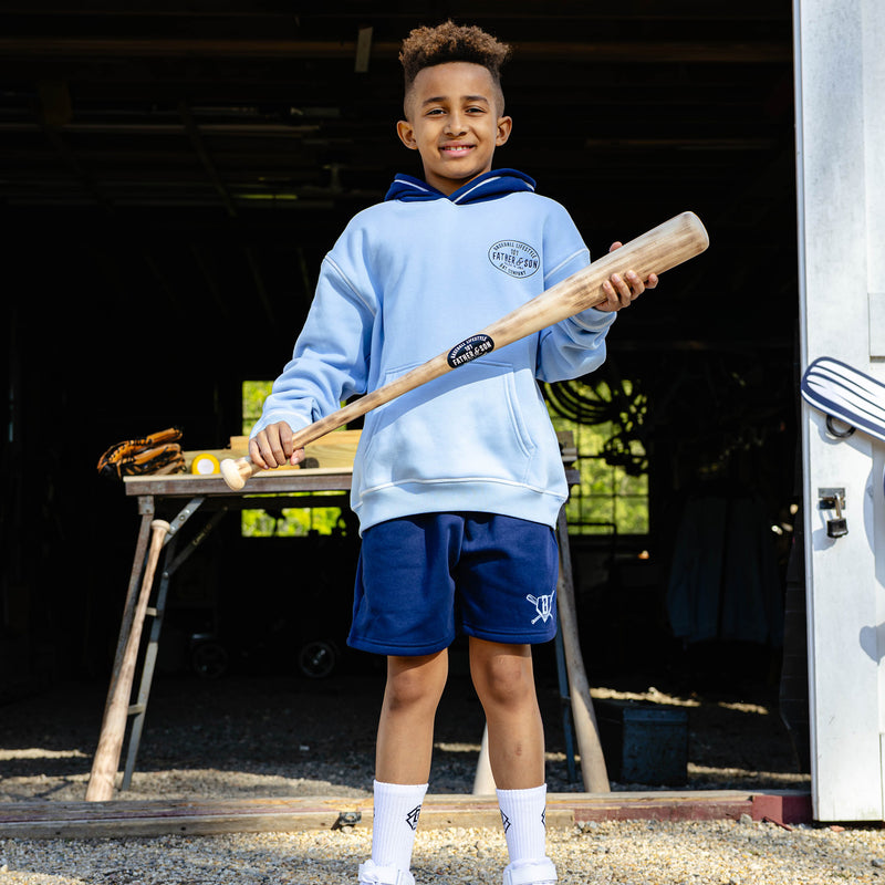 Young boy smiling and holding a wooden baseball bat while wearing the navy youth Heritage Diamond Sweatshort paired with a columbia blue Father & Son Bat Company hoodie and white BL101 crew socks.