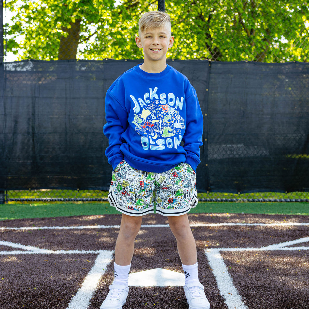 Smiling youth player at a baseball field wearing the royal blue Jackson Olson x BL101 Hometown Crewneck paired with the white Hometown Shorts