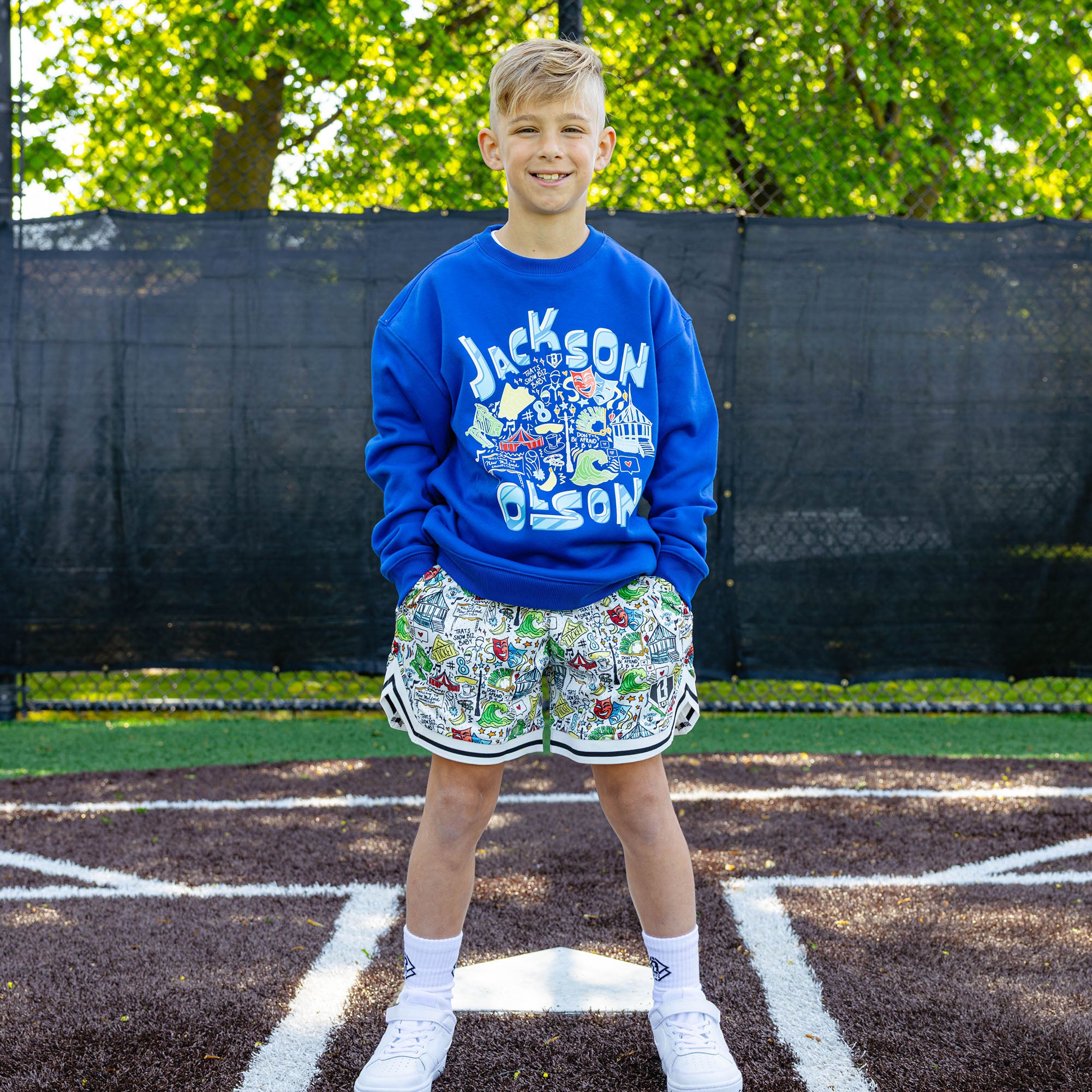 Smiling youth player at a baseball field wearing the royal blue Jackson Olson x BL101 Hometown Crewneck paired with the white Hometown Shorts