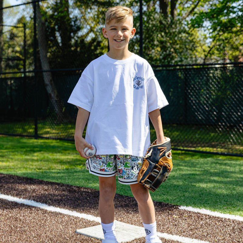 Youth player standing on a base holding a ball and glove, wearing the white Jackson Olson x BL101 relaxed tee with chest logo