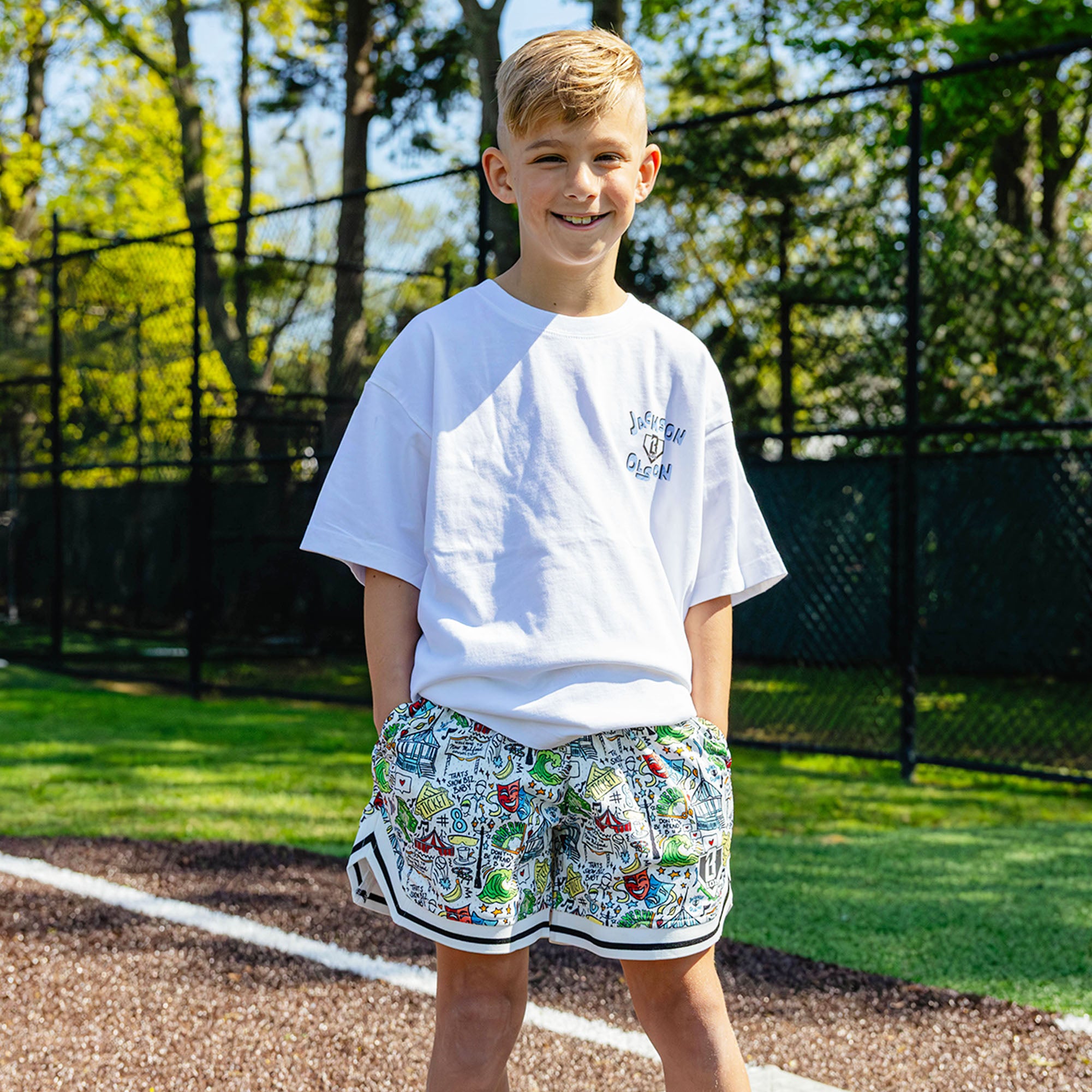 Smiling youth player on a baseball field wearing the white Jackson Olson x BL101 Hometown Shorts with all-over illustrated print paired with the white Jackson Olson Hometown Relaxed Tee