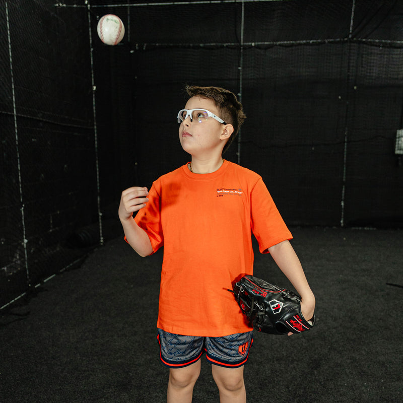Child in an orange shirt and black baseball glove preparing to catch a ball indoors.