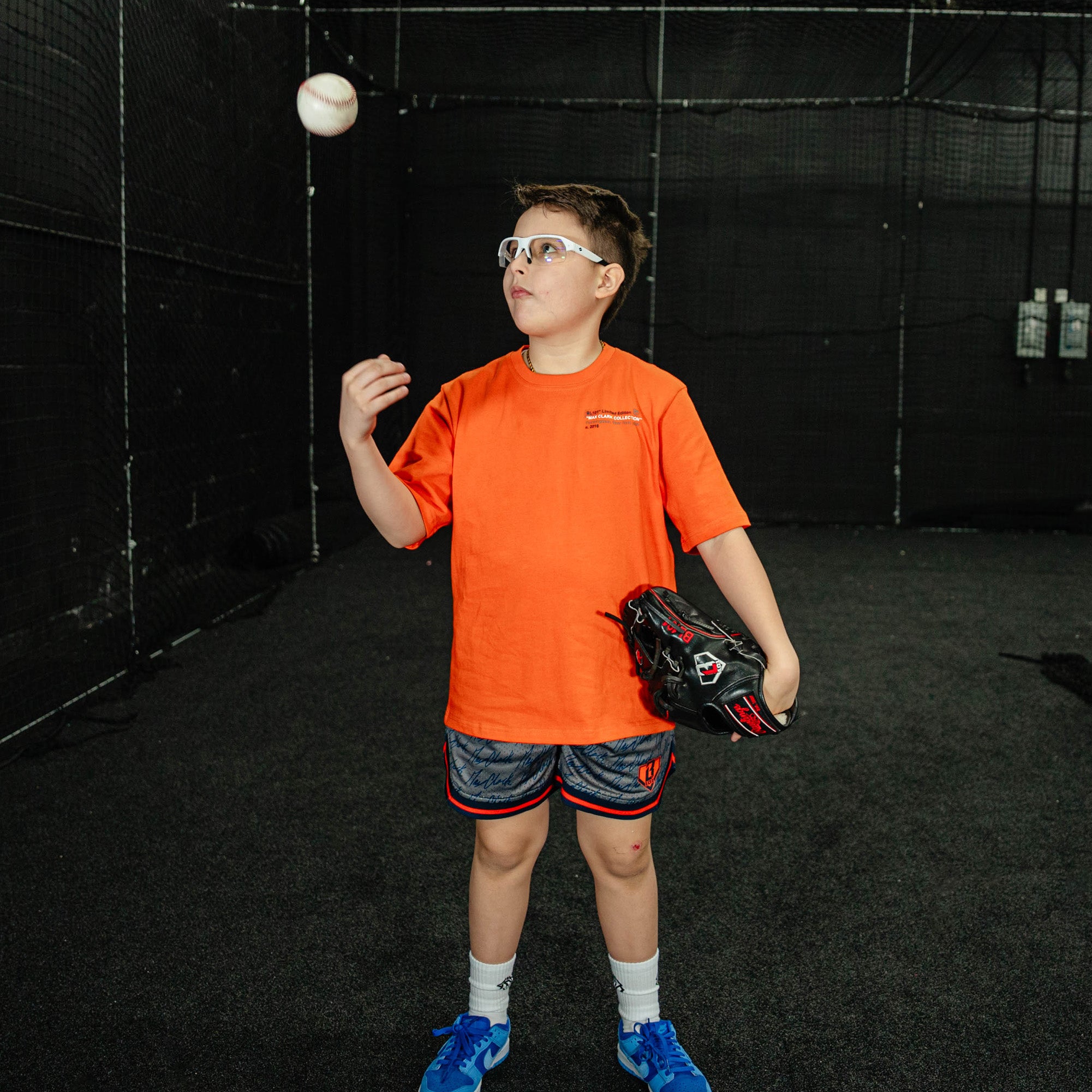 Child in an indoor batting cage wearing an orange shirt and blue shoes, preparing to catch a ball.