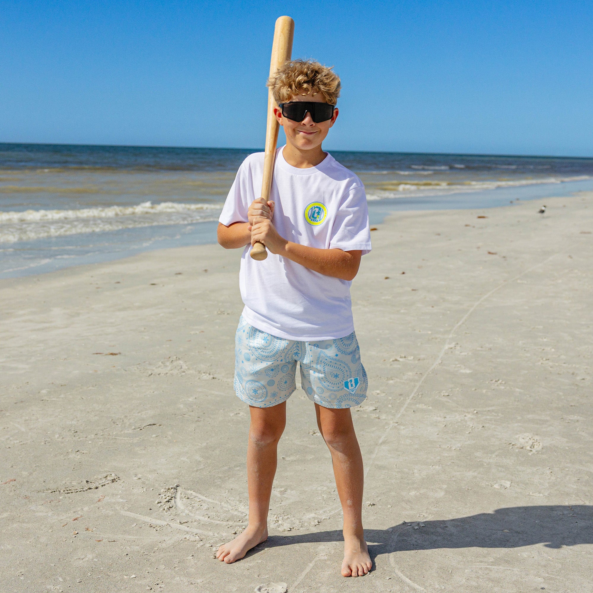 Young boy wearing BL101 blue paisley swim shorts and white tee, holding a baseball bat on the beach.
