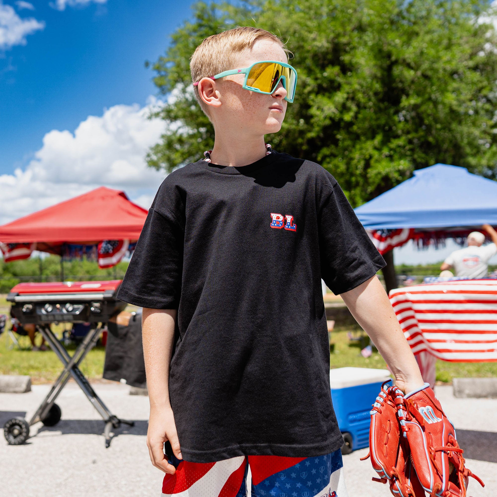 Front-facing youth male model wearing black Patriot Pitch tee with red white and blue shorts holding baseball glove with teal shield sunglasses at outdoor baseball event